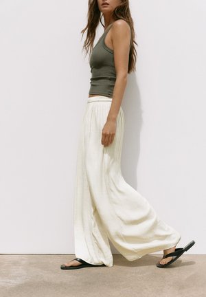 Femme portant un débardeur vert, un pantalon blanc ample et des sandales noires, marchant devant un fond clair uni.