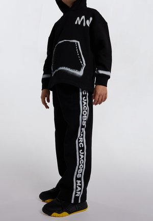 Dziecko w czarnej bluzie z kapturem z białym nadrukiem na kieszeni oraz czarnych spodniach dresowych z pionowym białym napisem "MARC JACOBS" po boku, czarne buty.