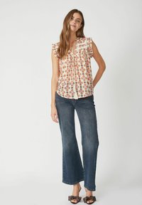 Top senza maniche in crema con un motivo geometrico arancione e marrone, con spalline arricciate e chiusura a fiocco sul collo. Abbinato a jeans svasati.