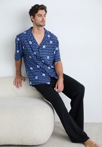Short-sleeve blue shirt with black and white repeating "HUGO" pattern, paired with black pants. Smooth fabric and button-up design.