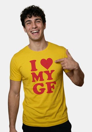 Hymyilevä nuori mies keltaisessa paidassa osoittaa punaista tekstiä, jossa lukee "I [heart] MY GF", yksinkertaisen harmaan taustan edessä.