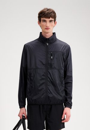 Νεαρός άνδρας που φοράει μαύρο ελαφρύ μπουφάν με φερμουάρ και μαύρο σορτς, κρατώντας το λουράκι μιας μαύρης τσάντας πάνω σε μονόχρωμο φόντο.