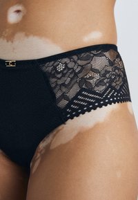 Hoogwaardige zwarte slips met kantdetails, bloemmotief, golvende randen en een kleine gouden accent op de tailleband.