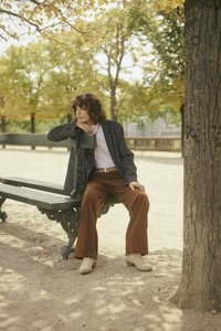 Mallin tumman teksturoidun takin päällä valkoinen paita, ruskeat samettihousut ja vaaleat nilkkurit, istuen vihreällä penkillä puistossa.