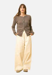 Brązowy sweter z dzianiny o fakturowanym wzorze, z sześcioma guzikami, w połączeniu z kremowymi spodniami o szerokich nogawkach i beżowym obuwiem.