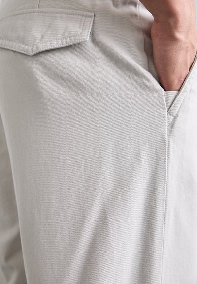 Person’s hand resting inside the front pocket of light beige pants with a back pocket flap visible.
