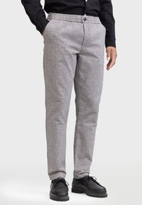 Man in grijze geruite broek met elastische taille en knop, gecombineerd met een zwart stoffen shirt en zwarte veterschoenen.