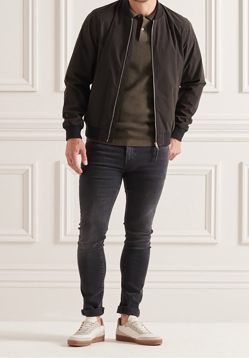 Homme portant une veste bomber noire, un pull polo vert olive, un jean slim foncé et des baskets blanches, debout à l'intérieur.