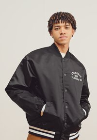 Chaqueta varsity de satén negro con detalles en blanco, que cuenta con un cierre de botones y un diseño bordado "Clase del 89" en el pecho.