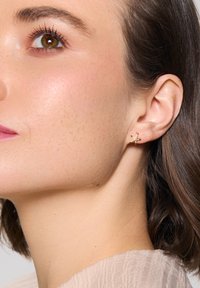 Złote kolczyki w kształcie gwiazdy morskiej z teksturowanym wykończeniem, z małymi kolorowymi akcentami. Lekka konstrukcja widoczna z bliska na uchu modelki.