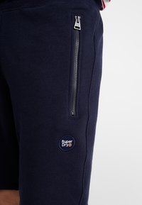Námořnické bavlněné šortky s bočním zipovým kapsou, s kulatou nášivkou loga "Super Dry" na dolní části nohy. Měkká textura a sportovní design.