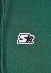 Tessuto di felpa verde con un logo ricamato con contorno bianco e dettagli a forma di stella nera sul lato destro.