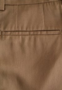 Pantalon marron avec une texture tissée lisse, doté d'une poche arrière unique et d'un motif de côtes diagonales subtil.