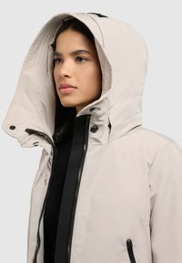 Jonge vrouw in een lichtbeige hoodie-jack met rits en zwarte binnenlaag, met haar gezicht naar links op een lichtgrijze achtergrond.