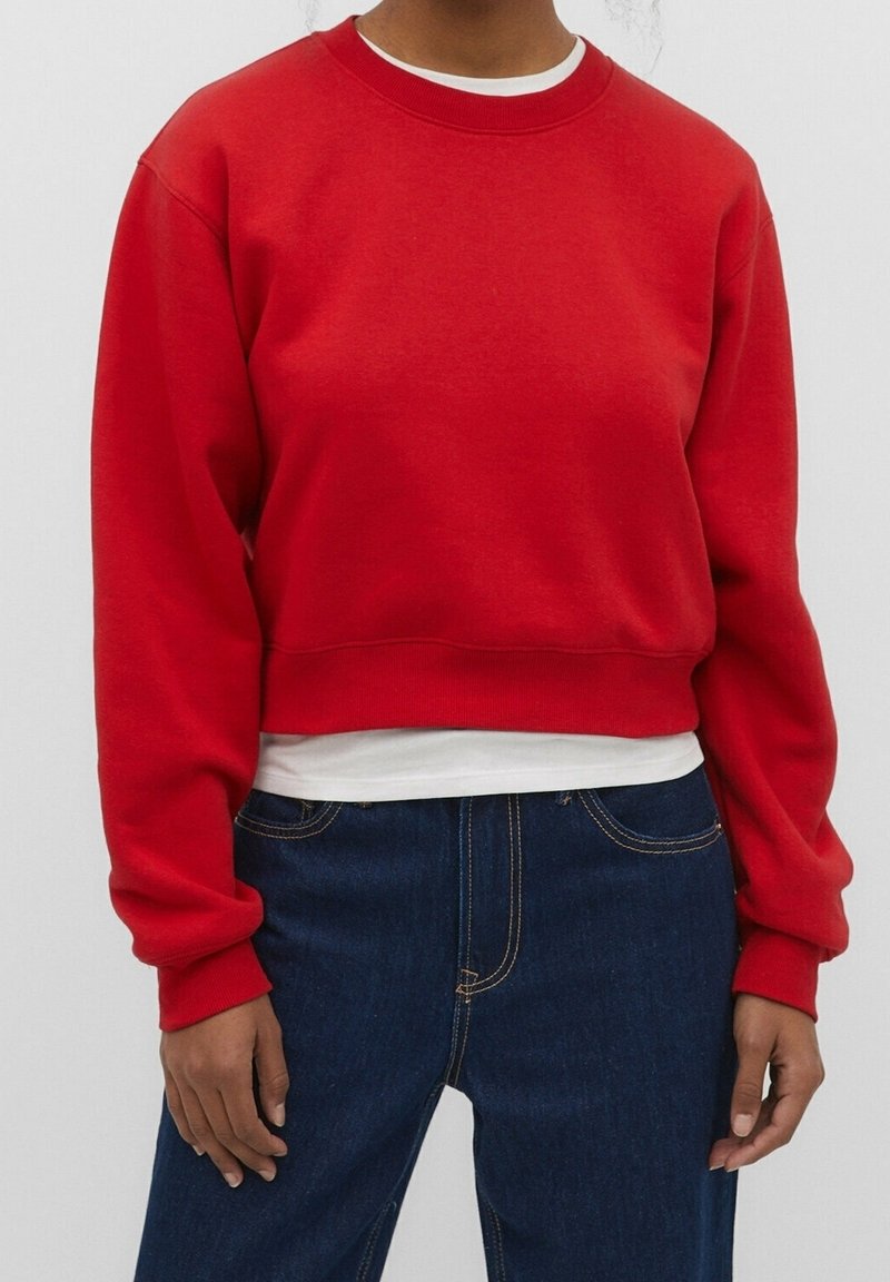 Personne portant un sweat court rouge à manches longues par-dessus une chemise blanche, associé à un jean bleu foncé sur un fond uni.