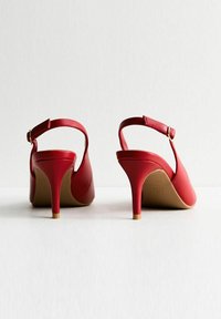 Röda slingback-hälar med en slät läderfinish, stängd tå och en tunn stilettklack. Har ett band med en guldbuckledetalj.