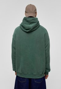 Groene katoenen hoodie met een textuur, een oversized capuchon, verlaagde schoudernaden en een geribbelde zoom, gecombineerd met een beige baseballpet.