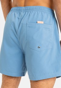 Light blue swim shorts featuring an elastic waistband, rear pocket with a metal grommet, and a subtle brand tag on the pocket.