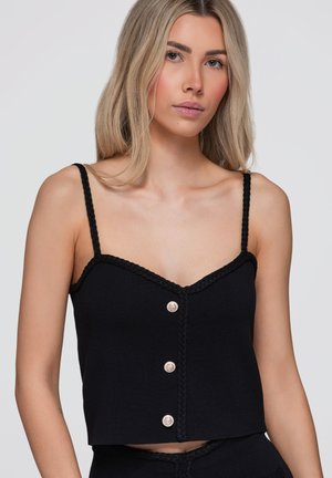 Jeune femme aux longs cheveux blonds portant un haut noir sans manches boutonné avec de fines bretelles tressées sur un fond uni.