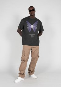 Mørkegrå t-shirt med et stort lilla sommerfugl grafik, kombineret med beige cargo-bukser med flere lommer og hvide sneakers.