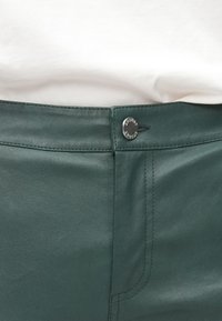 Donkergroene nepleren broek met een gladde textuur, voorzakstiksels en een metalen knoop met merkdetail.