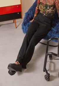 Pantalon noir taille haute, assorti à des chaussures à plateforme noires avec une bride à la cheville. L'arrière-plan présente une chaise texturée bleue.