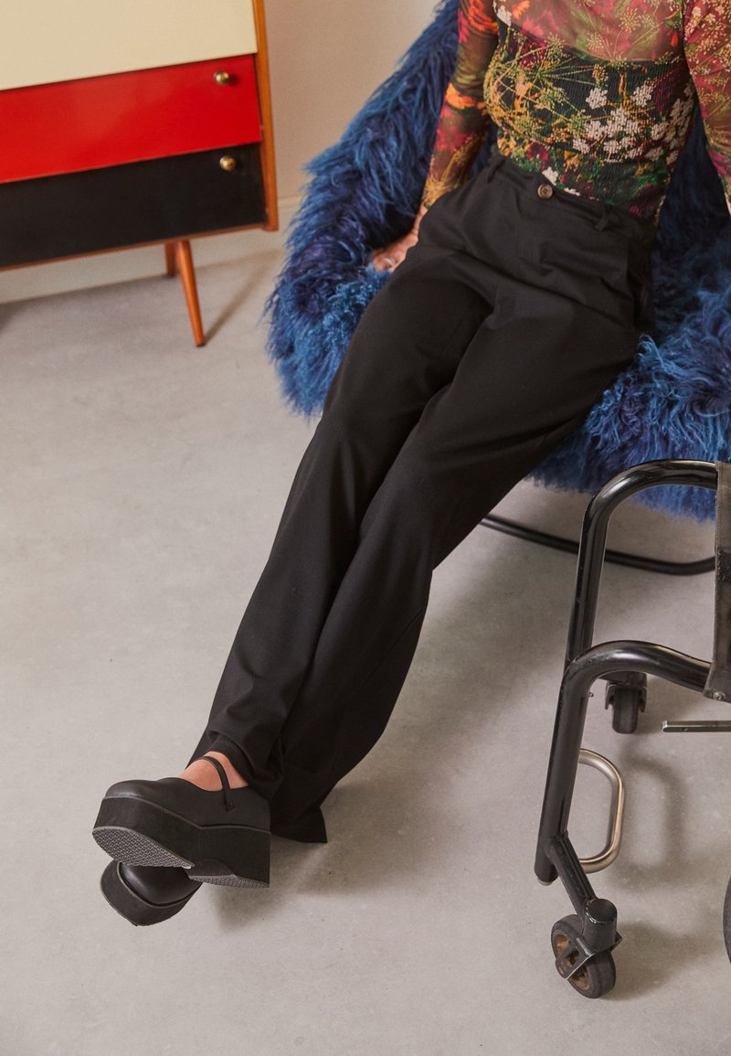 Pantalon noir taille haute, assorti à des chaussures à plateforme noires avec une bride à la cheville. L'arrière-plan présente une chaise texturée bleue.