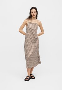 Γυναίκα φοράει ένα αμάνικο, μίντι φόρεμα με σχέδια και λεπτές τιράντες και μαύρα σανδάλια, στέκεται μπροστά σε ένα απλό λευκό φόντο.