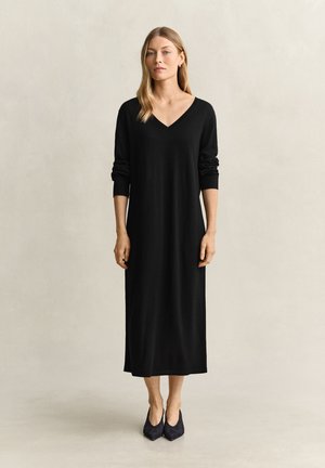 Femme aux cheveux blonds et raides portant une longue robe noire à col en V avec des manches longues et des chaussures noires pointues, debout devant un fond uni.