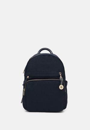 Sac à dos noir avec une poche zippée à l'avant et un petit charm rond doré sur la tirette du zip, sur un fond clair uni.