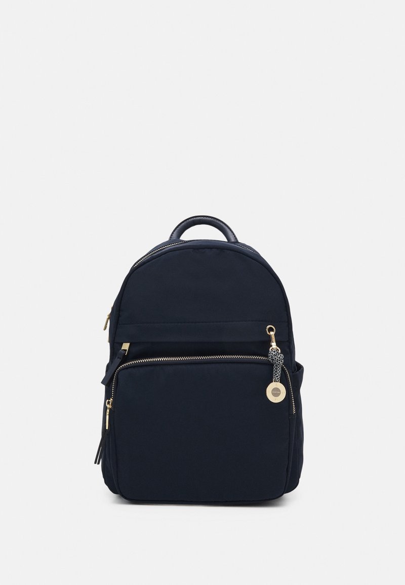 Sac à dos noir avec une poche zippée à l'avant et un petit charm rond doré sur la tirette du zip, sur un fond clair uni.