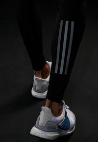 Leggings neri con design a tre strisce argentee, abbinati a sneaker grigie e bianche con una tomaia testurizzata e dettagli blu.