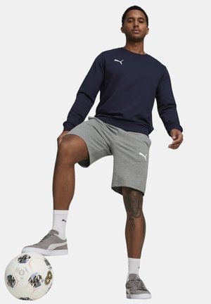 Felpa blu navy con logo bianco, pantaloni corti grigi e sneakers grigie. In piedi accanto a un pallone da calcio, presenta un tatuaggio visibile sulla gamba.