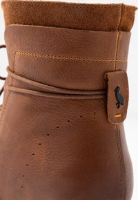 Bottine en cuir marron avec un col en daim texturé, des coutures décoratives et une étiquette sur le côté avec un emblème d'oiseau.