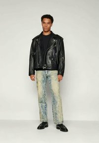 Jonge man in een zwart leren motorjack, zwart shirt, licht versleten jeans en zwarte leren schoenen, staand tegen een effen achtergrond.