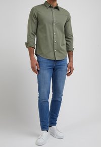 Man in een olijfgroene button-up shirt met opgerolde mouwen, blauwe slim-fit jeans en witte sneakers, staande tegen een effen achtergrond.