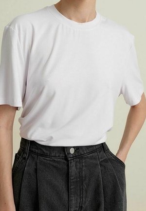 Persoon draagt een effen wit T-shirt met korte mouwen, ingestopt in een hooggetailleerde, zwarte geplooide broek, met handen in de zakken.