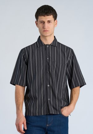 Giovane uomo con capelli corti e scuri indossa una camicia nera a maniche corte con righe verticali bianche e jeans blu, una mano in tasca.
