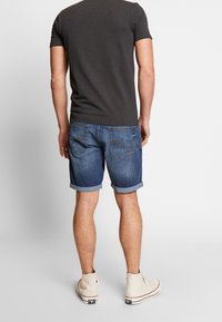 T-shirt gris foncé pour homme, associé à un short en jean bleu retroussé. Détails de couture remarquables sur les poches arrière, et chaussures décontractées de couleur claire.