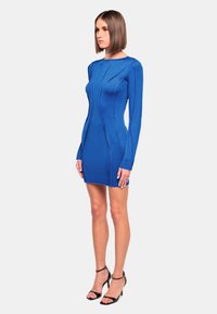 Kurzes blaues Bodycon-Kleid mit langen Ärmeln, das vertikale Nähte und eine enganliegende Silhouette aufweist. Tragen Sie dazu schwarze, gestreifte High Heels.
