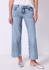 Vaaleansinisten leveälahkeisten farkkujen, joissa on risat helmat, viisi taskua ja nappikiinnitys vyötäröllä, päällä hopeiset ballerinat.