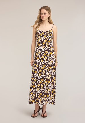 Joven mujer con vestido maxi marrón sin mangas con estampado floral amarillo y sandalias negras de tacón alto, de pie sobre un fondo liso.