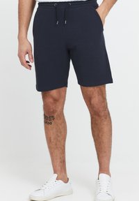 Mørkeblå bomulds shorts med elastisk talje, snøring og to sidelommer; slutter lige over knæet. Hvide sneakers.