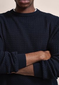 Marinblauer gestrickter Pullover mit einem strukturierten Muster, Rundhalsausschnitt und gerippten Bündchen; der Stoff wirkt weich und dick. Die Arme des Models sind crossed.