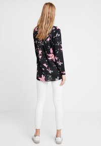 Czarna bluzka z długim rękawem w różowy kwiatowy wzór, okrągły dekolt i lekko luźny krój, zestawiona z białymi obcisłymi spodniami i szarymi butami.