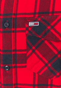 Rood en zwart geruite flanellen shirt met een knoopsluiting aan de voorkant, een zwarte zak en een geborduurd logo op de borst. Textuurstof.