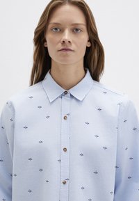 Light blue button-up shirt with a collar, featuring small embroidered floral patterns in dark blue. Smooth fabric texture and regular fit.