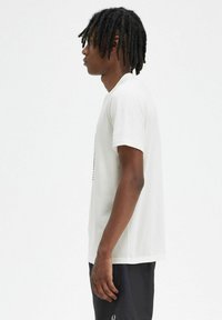Camiseta de algodón blanca con mangas cortas y cuello redondo, que presenta un diseño estampado en la parte delantera y un corte relajado. Llevada con pantalones cortos oscuros.
