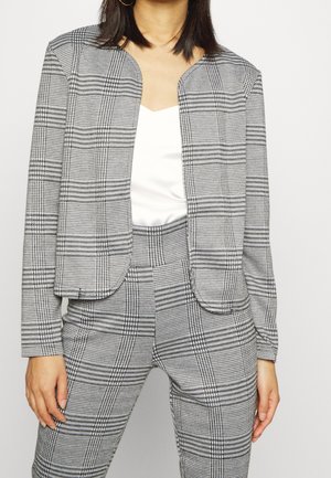 Veste de costume à carreaux gris avec un devant arrondi, des manches longues et sans fermeture. Associée à un haut blanc et à un pantalon à carreaux assorti.
