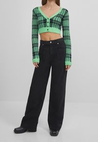 Zielono-czarny krótki kardigan w kratę z długimi rękawami i zapięciem na guziki z przodu, zestawiony z czarnymi, szerokimi jeansami z wysokim stanem oraz czarnymi butami.